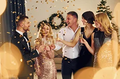 Grupa pięciu osób w strojach wieczorowych świętuje przy szampanie. Uśmiechają się, otoczeni świątecznymi lampkami i dekoracjami, emanując radością i świątecznym nastrojem.