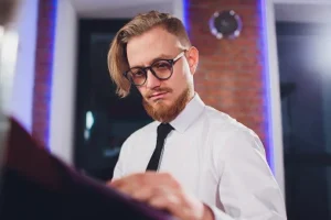 Mężczyzna w okularach, z rudą brodą i gładko zaczesanymi do tyłu włosami ogląda tkaninę w nowoczesnym otoczeniu. Ma na sobie białą koszulę i czarny krawat, co podkreśla jego skupienie i styl.