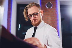 Mężczyzna w okularach, z rudą brodą i gładko zaczesanymi do tyłu włosami ogląda tkaninę w nowoczesnym otoczeniu. Ma na sobie białą koszulę i czarny krawat, co podkreśla jego skupienie i styl.