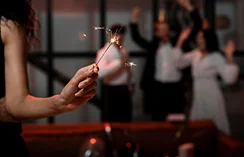 Zbliżenie dłoni trzymającej zapaloną sztuczną ogniem na słabo oświetlonej imprezie. Na zamazanym tle widać trzy osoby w świątecznych strojach i koronach, świętujące.