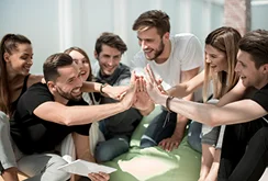 Grupa siedmiu osób siedzi w jasnym pomieszczeniu, uśmiecha się i przybija sobie piątki, wyrażając poczucie pracy zespołowej i radości. Są ubrani swobodnie i wyglądają na zadowolonych.